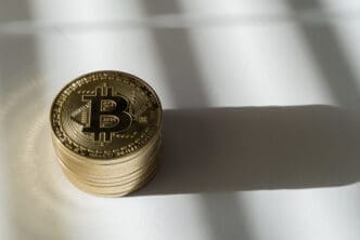 A high-angle view of a neat stack of golden Bitcoin coins on a plain white surface, casting a distinct, elongated shadow.