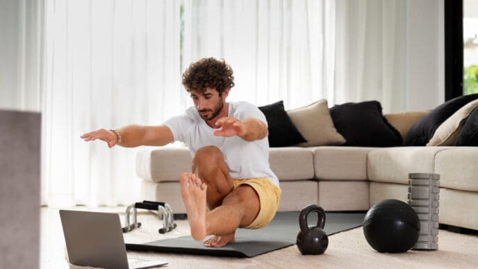A man performs a single-leg squat exercise on a yoga mat in a bright living room, with a laptop in front of him and various fitness equipment like a kettlebell and medicine ball scattered around.