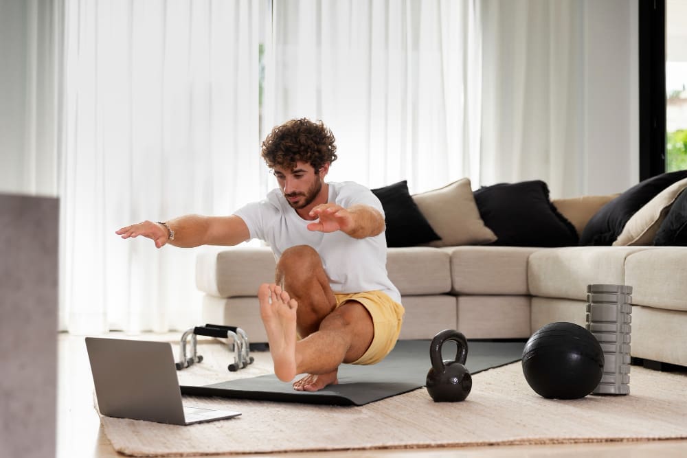 A man performs a single-leg squat exercise on a yoga mat in a bright living room, with a laptop in front of him and various fitness equipment like a kettlebell and medicine ball scattered around.