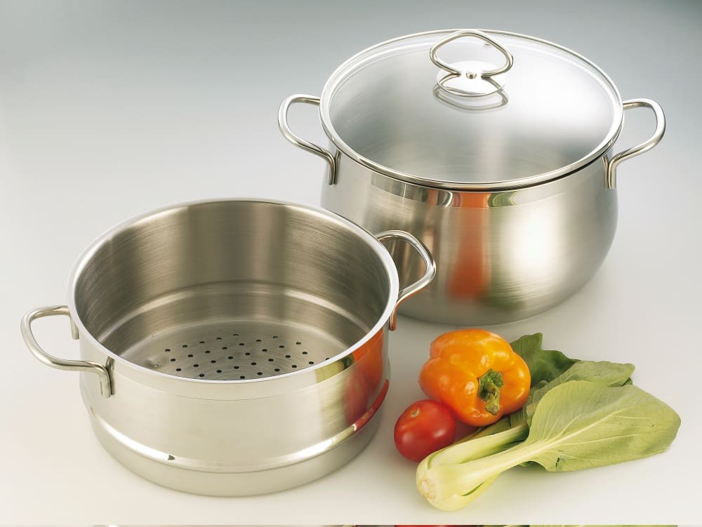 A set of stainless steel cooking pots, one with a steamer insert, next to fresh vegetables like peppers, tomatoes, and bok choy.