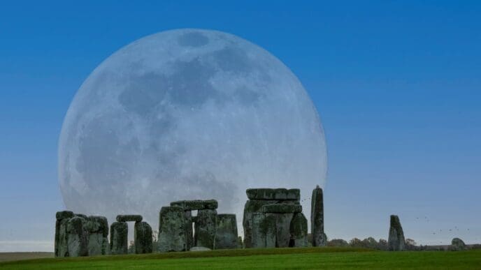 A large, luminous full moon rising behind the ancient stone circle of Stonehenge, with green grass in the foreground under a clear blue sky.