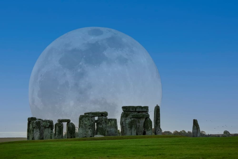 A large, luminous full moon rising behind the ancient stone circle of Stonehenge, with green grass in the foreground under a clear blue sky.