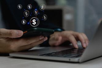 Close-up of hands holding a smartphone displaying a glowing dollar sign and various floating dollar coin icons, while simultaneously typing on a laptop keyboard, symbolizing digital financial transactions or a fintech concept.