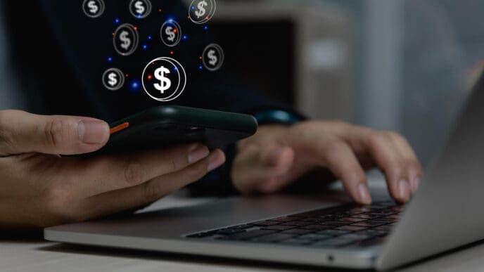 Close-up of hands holding a smartphone displaying a glowing dollar sign and various floating dollar coin icons, while simultaneously typing on a laptop keyboard, symbolizing digital financial transactions or a fintech concept.