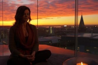 A woman meditating or practicing yoga in a lotus position on a rooftop terrace, silhouetted against a vibrant sunset over a city.