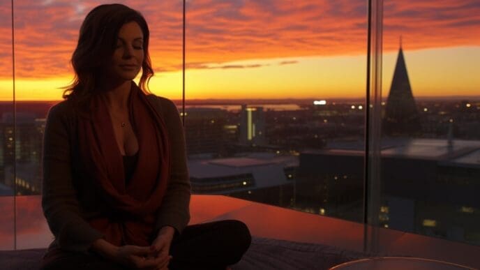 A woman meditating or practicing yoga in a lotus position on a rooftop terrace, silhouetted against a vibrant sunset over a city.