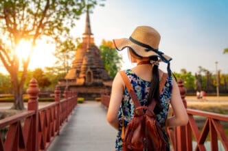 Rear view of a woman in a straw hat and floral dress walking on a wooden bridge towards a historic stupa, with a sunset in Sukhothai Historical Park, Thailand.