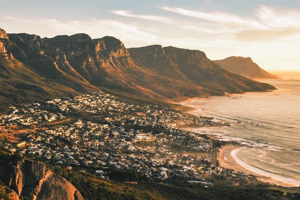 Aerial view of Camps Bay, Cape Town, with mountains, coastline, and residential areas illuminated by sunset.