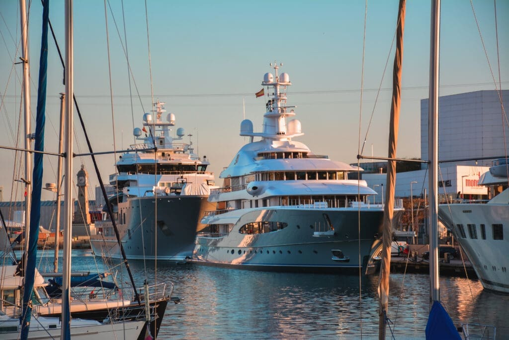 Two large, luxurious superyachts moored in a harbor, surrounded by smaller sailboats and city buildings in the background at sunset.