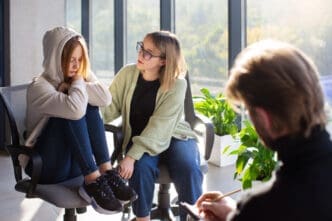 A young woman in a hoodie sits with her head down, receiving comfort from another woman in glasses, while a male therapist (seen from behind) writes notes, depicting a psychotherapy session.
