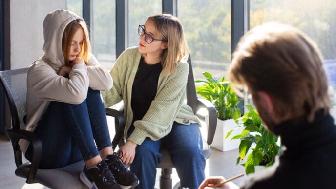 A young woman in a hoodie sits with her head down, receiving comfort from another woman in glasses, while a male therapist (seen from behind) writes notes, depicting a psychotherapy session.