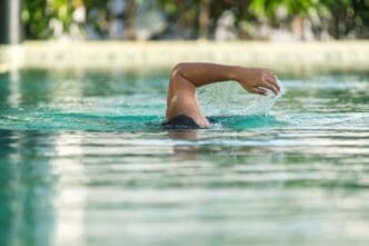 A person's arm breaking the surface of clear blue pool water during a swim stroke, with blurred greenery in the background.