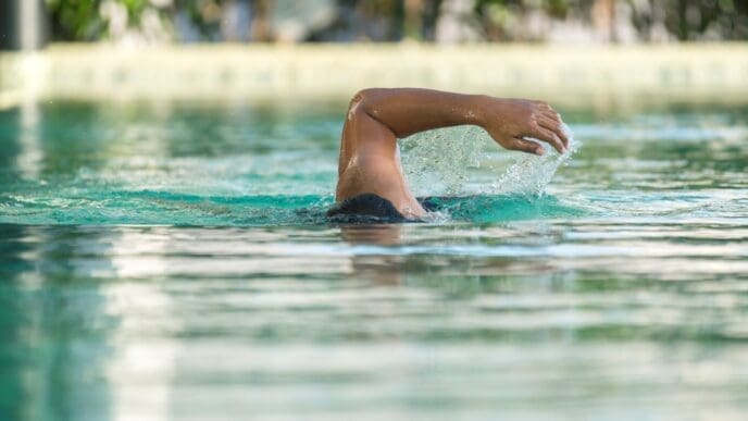 A person's arm breaking the surface of clear blue pool water during a swim stroke, with blurred greenery in the background.