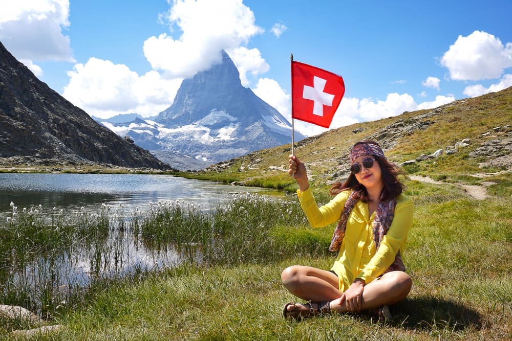 A happy Asian woman sitting by an alpine lake, holding a Swiss flag, with the Matterhorn mountain in the background.