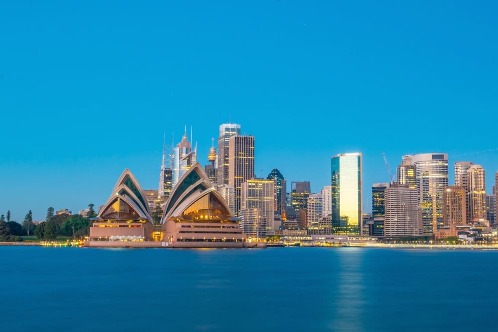 The Sydney Opera House and city skyline at dusk, reflected in the still water of the harbor.