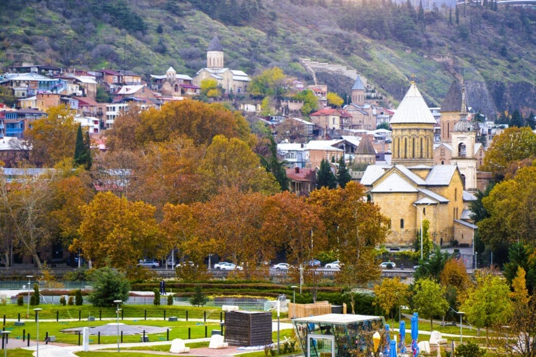Panoramic view of Tbilisi, Georgia, with the Rike Park in the foreground, and historic churches and buildings on the hillside, surrounded by autumn trees.