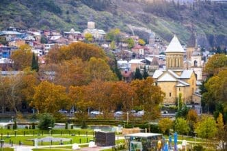 Panoramic view of Tbilisi, Georgia, with the Rike Park in the foreground, and historic churches and buildings on the hillside, surrounded by autumn trees.