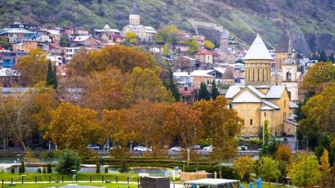 Panoramic view of Tbilisi, Georgia, with the Rike Park in the foreground, and historic churches and buildings on the hillside, surrounded by autumn trees.