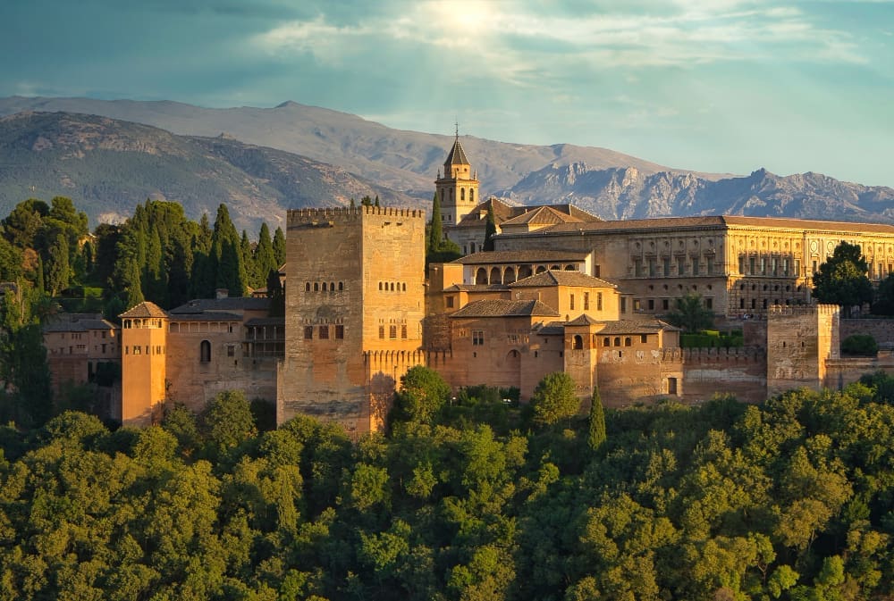 The magnificent Alhambra palace and fortress complex, with its warm-toned walls, towers, and diverse architecture, nestled amidst lush green trees on a hill, with distant mountains under a partly cloudy sky at sunset.