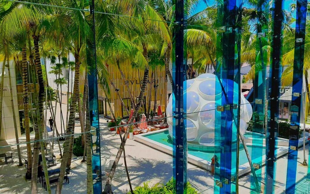 An indoor-outdoor view of the Miami Design District's Palm Court, showing a public space with a white, bubble-like "Fly's Eye Dome" structure and a pool, surrounded by palm trees and modern buildings, as seen through a blue-tinted glass wall.