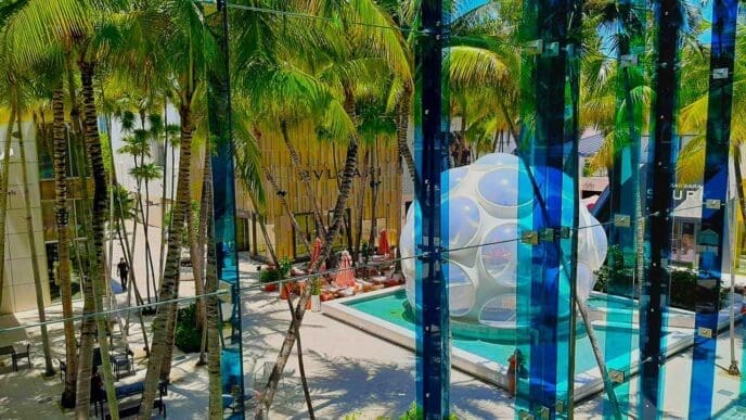 An indoor-outdoor view of the Miami Design District's Palm Court, showing a public space with a white, bubble-like "Fly's Eye Dome" structure and a pool, surrounded by palm trees and modern buildings, as seen through a blue-tinted glass wall.