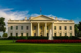 The North Portico of the White House is shown in a wide shot, with its white columns, fountain, manicured green lawn, and a large bed of vibrant red flowers, under a clear blue sky.