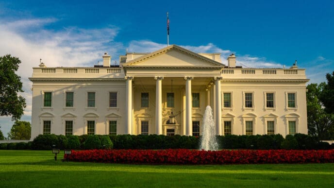 The North Portico of the White House is shown in a wide shot, with its white columns, fountain, manicured green lawn, and a large bed of vibrant red flowers, under a clear blue sky.