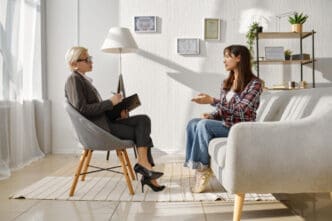 A female therapist with short blonde hair and glasses taking notes while her young female client, with dark hair, speaks and gestures during a therapy session.