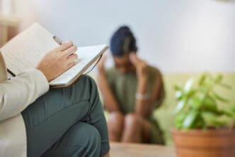 A therapist taking notes during a session, with a distressed patient blurred in the background, symbolizing mental health support.