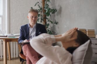 A man, possibly a therapist, looking at a person lying on a couch with their face covered, suggesting emotional distress.
