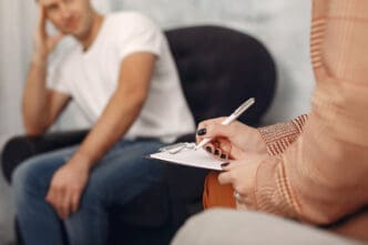 A therapist, seen from the shoulder down, writing on a clipboard, with a blurred male patient in a white t-shirt and jeans sitting in the background, representing a therapy session.