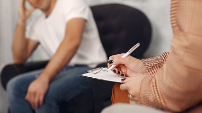 A therapist, seen from the shoulder down, writing on a clipboard, with a blurred male patient in a white t-shirt and jeans sitting in the background, representing a therapy session.