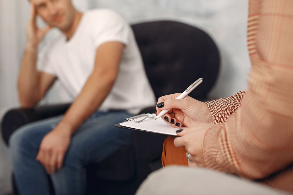 A therapist, seen from the shoulder down, writing on a clipboard, with a blurred male patient in a white t-shirt and jeans sitting in the background, representing a therapy session.