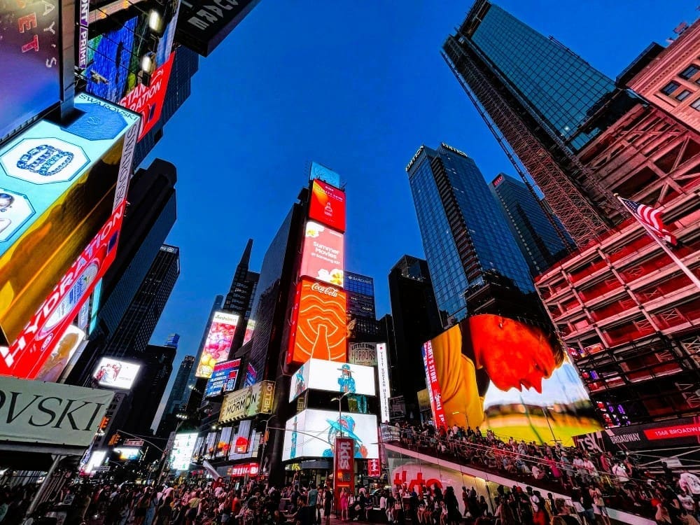 A low-angle, wide shot of the bustling Times Square in New York City at dusk, with a dense cluster of towering skyscrapers and numerous brightly lit digital billboards and advertisements under a deep blue sky.