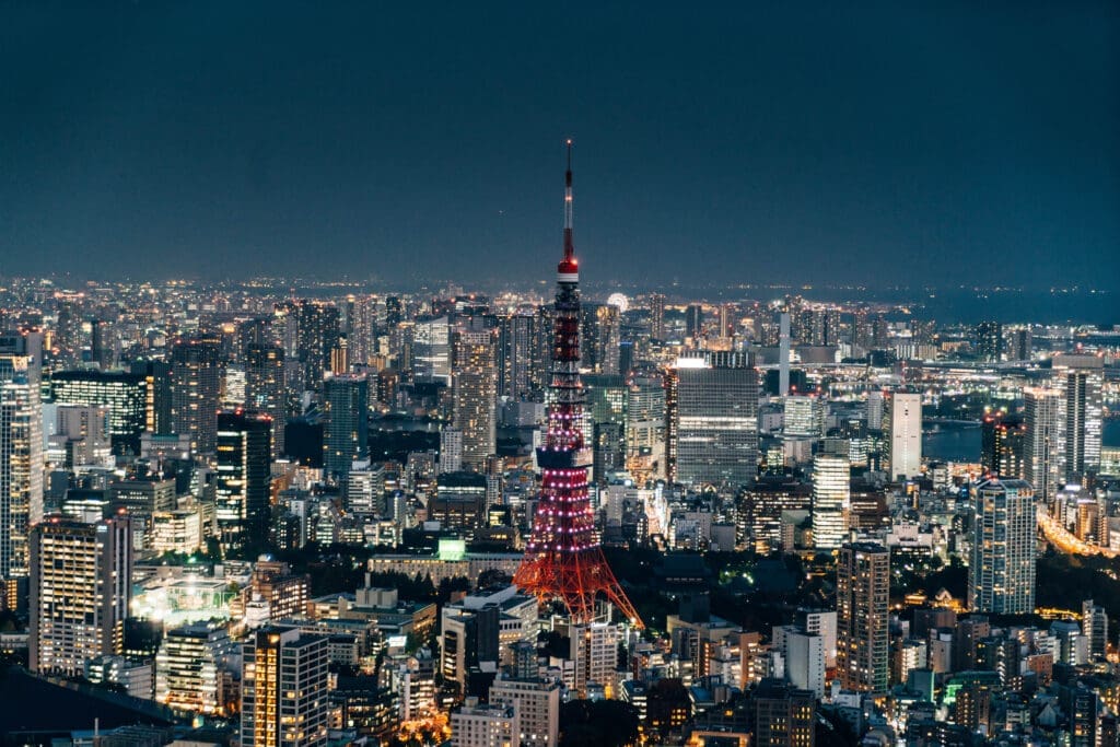 A high-angle aerial view of the Tokyo skyline at night, with the illuminated red and white Tokyo Tower rising prominently above thousands of city lights.