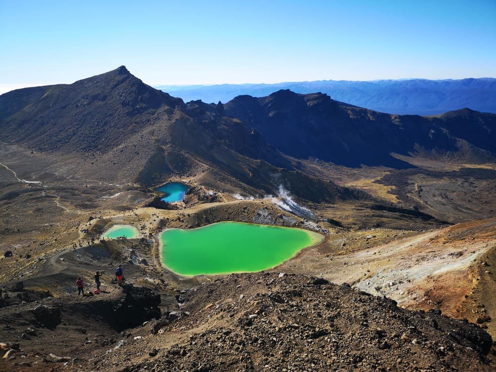 A high-angle panoramic view of a volcanic landscape in Tongariro National Park, featuring three vibrant emerald and blue crater lakes, steaming vents, rugged brown and black terrain, and distant mountains under a clear blue sky.
