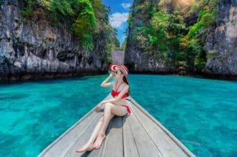 A beautiful girl in a red bikini and striped sun hat sits on the bow of a wooden longtail boat, cruising on turquoise waters surrounded by lush, towering limestone cliffs in Thailand.