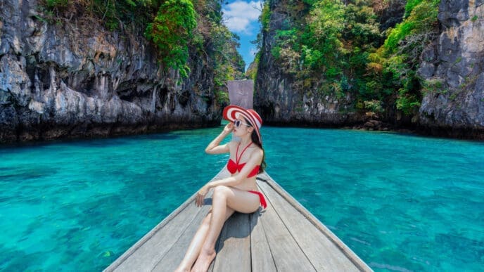 A beautiful girl in a red bikini and striped sun hat sits on the bow of a wooden longtail boat, cruising on turquoise waters surrounded by lush, towering limestone cliffs in Thailand.