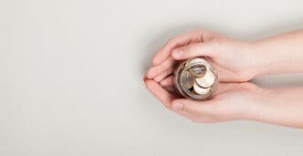 Two hands gently hold a small glass jar filled with coins, symbolizing saving, investing, or trust-based philanthropy, against a plain light background.