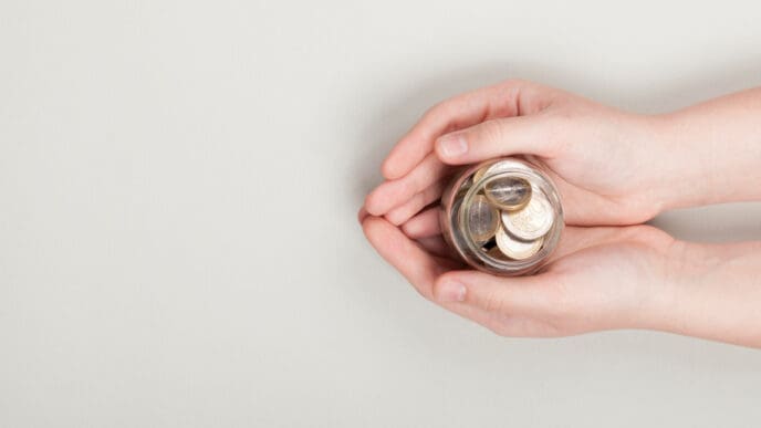Two hands gently hold a small glass jar filled with coins, symbolizing saving, investing, or trust-based philanthropy, against a plain light background.