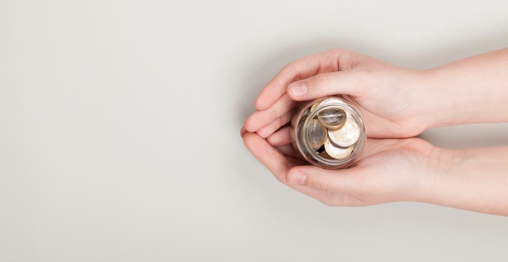 Two hands gently hold a small glass jar filled with coins, symbolizing saving, investing, or trust-based philanthropy, against a plain light background.