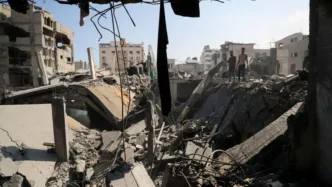 A wide view of a devastated urban area, showing extensive rubble, twisted rebar, and collapsed buildings in the aftermath of conflict, with two young men standing on a pile of debris in the distance.