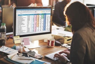 A person working on a computer screen displaying a financial spreadsheet, with a calculator and office supplies on the desk.