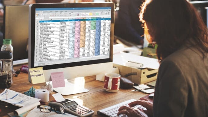 A person working on a computer screen displaying a financial spreadsheet, with a calculator and office supplies on the desk.