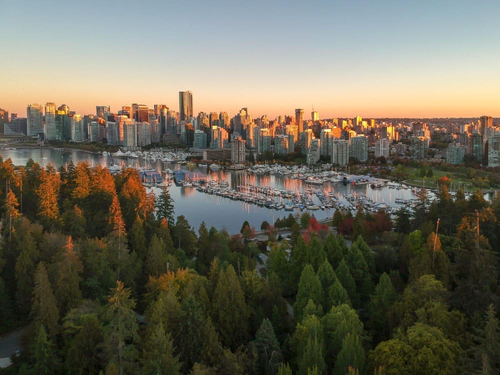 A panoramic aerial view of the Vancouver downtown skyline with tall buildings glowing in the golden light of dusk, overlooking a marina filled with boats, surrounded by dense green trees and a body of water.
