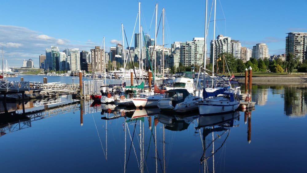 A marina filled with various sailboats and yachts, their masts reflected in the calm, deep blue water, with the modern skyline of Vancouver city buildings in the background under a clear blue sky.