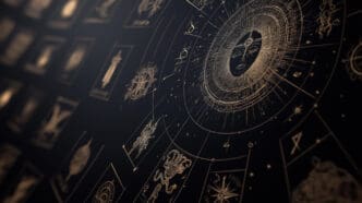 An ornate and detailed digital illustration of a glowing golden astrological chart with a sun or face symbol at its center, surrounded by various planetary glyphs and zodiac symbols, set against a dark background with blurred tarot-like cards in the foreground.