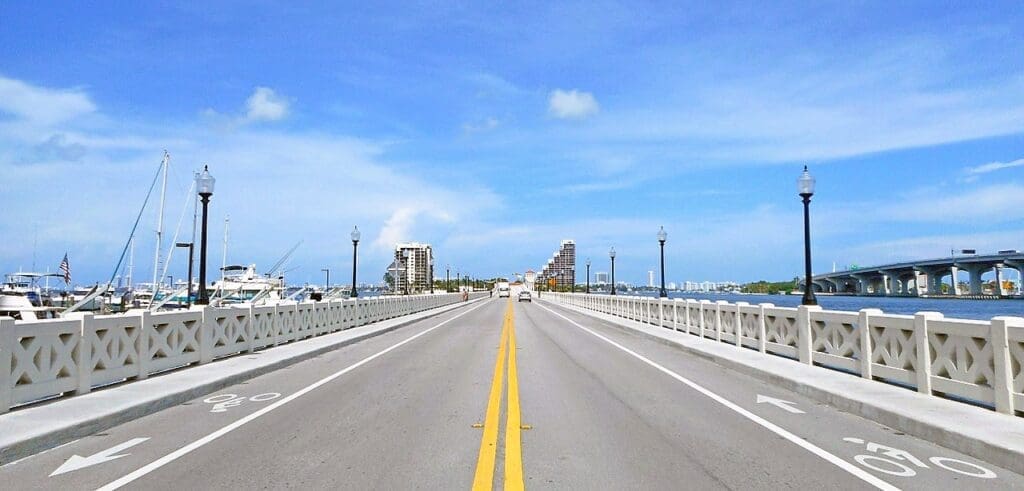 The Venetian Causeway stretching across blue water with a clear road, marina, and city skyline in the distance under a blue sky.