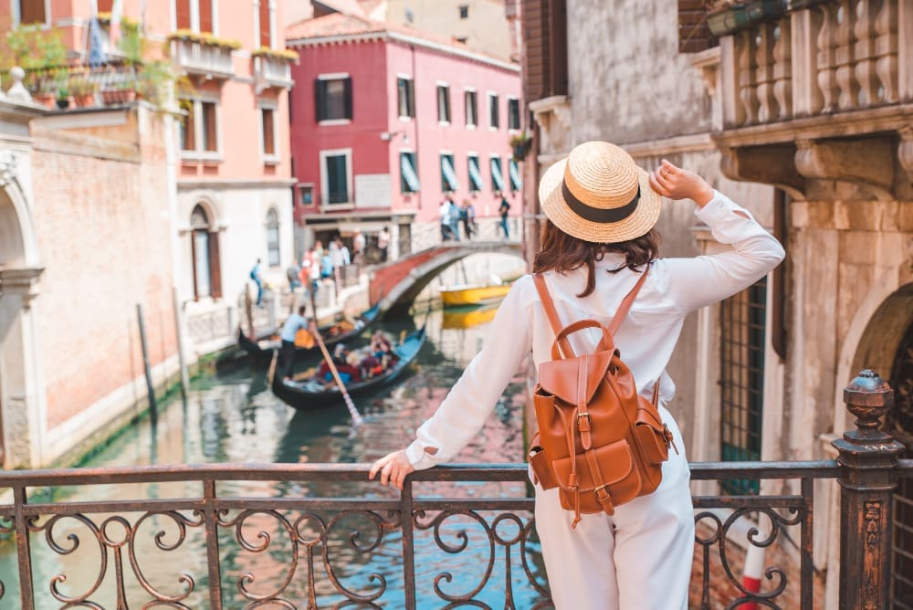 A woman in a hat and backpack looking out over a Venetian canal with gondolas and historic buildings.