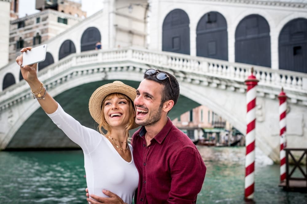 A happy couple taking a selfie in front of the iconic Rialto Bridge in Venice, Italy.